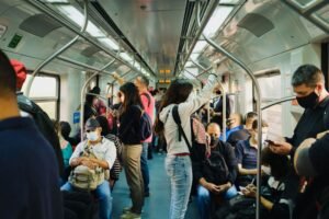 Crowded interior of a metro train with seated and standing passengers, most wearing face masks, during daytime. The scene reflects public transport usage with a diverse group of commuters holding onto handrails or using mobile phones.