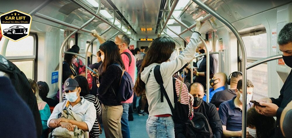 Crowded interior of a metro train with seated and standing passengers, most wearing face masks, during daytime. The scene reflects public transport usage with a diverse group of commuters holding onto handrails or using mobile phones. cheap car lift Dubai to Abu Dhabi